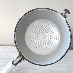 May include: A vintage white enamelware sieve with a black rim and handle. The sieve features a perforated base for draining liquids. A classic kitchen utensil for straining pasta, vegetables, or other foods.