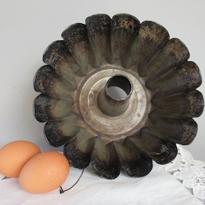 May include: A vintage, fluted metal bundt pan with a central tube, showing signs of wear and age. The pan has a dark, aged appearance with a flower-like design. Two brown eggs are next to the pan.