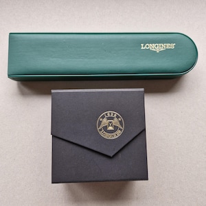 May include: A green rectangular watch case and a black watch box. The watch case has the word "LONGINES" printed in gold. The black box has a gold emblem with the words "1832 LONGINES" and an hourglass design.