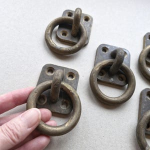 May include: Several antique brass ring pulls with rectangular backplates. The ring pulls are a warm, aged brass color, and the backplates have a darker, tarnished appearance. The hardware is likely for furniture or decorative use.