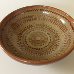 Może przedstawiać: Okrągła ceramiczna miska z jasnobeżowym wnętrzem i ciemnobrązową obwódką. Wnętrze zdobią koncentryczne okręgi i zygzakowate wzory w kolorze brązowym. Miska ma rustykalny, ręcznie robiony wygląd.