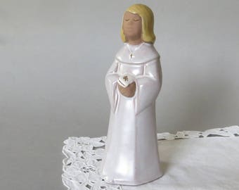 May include: A ceramic figurine of a person in a white robe with a cross pendant and closed eyes. The figurine has blonde hair and is holding a small, decorated object. The figure is standing on a white lace tablecloth.