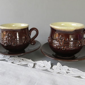 Puede incluir: Dos tazas de café de cerámica marrón con platillos a juego. Las tazas tienen un diseño decorativo en el lateral y un borde beige claro. Los platillos son de color marrón liso. Las tazas tienen un asa curva.