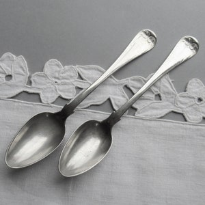 May include: Two vintage silver spoons with decorative floral patterns on the handles, placed on a white floral-edged cloth. The spoons have a classic, elegant design, suitable for serving or display. The spoons are set against a grey background.
