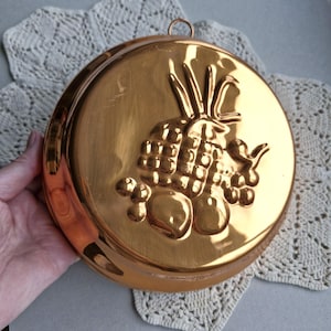 May include: A circular, copper-coloured metal mould with a fruit design. The embossed design features a pineapple, grapes, and other fruits. The mould has a loop for hanging and is held by a hand.