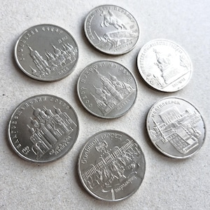 May include: A collection of silver coins featuring embossed architectural designs and Cyrillic text. The coins depict buildings, monuments, and historical figures. The coins are round and have a metallic sheen.