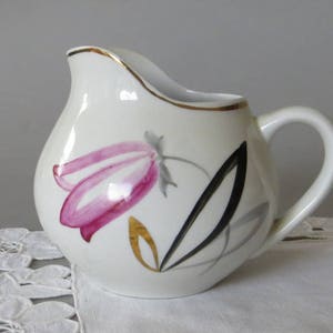 May include: A white ceramic creamer jug featuring a hand-painted pink bell-shaped flower with grey and gold leaf accents. The jug has a curved handle, a pouring spout, and a thin gold rim along the top edge of the vessel.