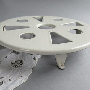 May include: White metal trivet with a circular base and three legs. The base has a decorative design with cutouts in the shape of circles and triangles.