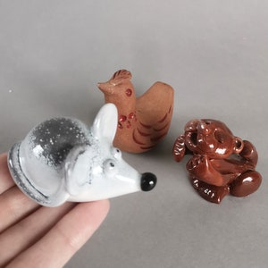 以下が含まれることがあります： 3つのセラミック動物フィギュア：グレーと白のネズミ、茶色の鶏、茶色のクマ。ネズミは黒い鼻をしており、鶏は赤いアクセントがあります。クマは座った姿勢です。