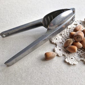 Puede incluir: Un cascanueces plateado vintage con mangos largos, abierto y listo para romper nueces. Varias avellanas marrones están esparcidas sobre un tapete de encaje blanco, lo que sugiere un tema rústico y otoñal. La herramienta es un clásico de cocina.
