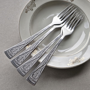 May include: Four silver forks with decorative, engraved handles are arranged on a white plate with a gold floral pattern. The forks are positioned diagonally, highlighting their intricate designs. The plate has a gold edge.