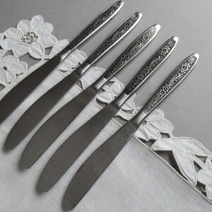 May include: Five silver dinner knives with floral designs on the handles. The knives are arranged in a row on a white lace tablecloth.