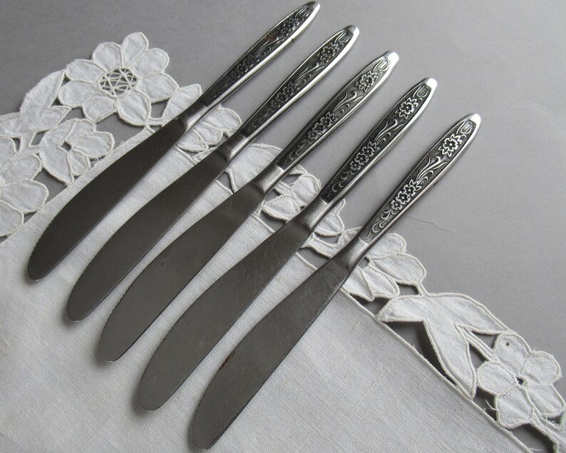 May include: Five silver dinner knives with floral designs on the handles. The knives are arranged in a row on a white lace tablecloth.