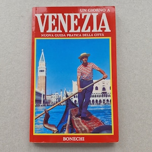 Könnte beinhalten: Ein roter Reiseführer mit dem Titel "Venezia" und dem Text "Un Giorno a Venezia" und "Nuova Guida Pratica Della Citta". Das Cover zeigt einen Gondoliere in Venedig, der ein gestreiftes Hemd und eine blaue Hose trägt und eine Gondel steuert.
