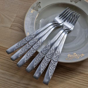 Peut inclure: Six fourchettes en argent avec des motifs de roses et de feuilles sur les manches sont disposées sur une assiette décorative. L'assiette est grise clair avec des accents floraux dorés. Sur une surface en bois.