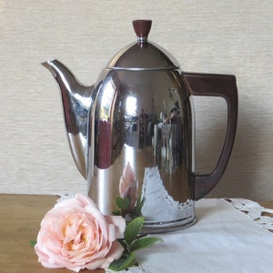 May include: A vintage chrome coffee pot with a dark brown handle and lid finial. The pot reflects the surrounding environment. A pink rose and white lace doily are in the foreground. The pot is a classic kitchenware item.