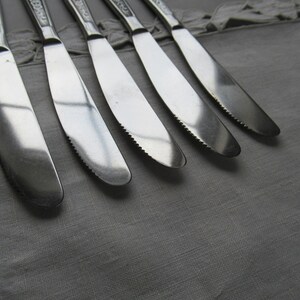May include: Six silver dinner knives with ornate handles are laid out on a white tablecloth. The knives are sharp and have a polished finish.