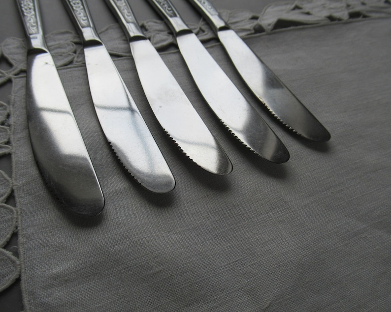 May include: Six silver dinner knives with ornate handles are laid out on a white tablecloth. The knives are sharp and have a polished finish.