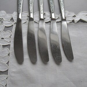 May include: Five silver butter knives with ornate handles lay on a white lace tablecloth.