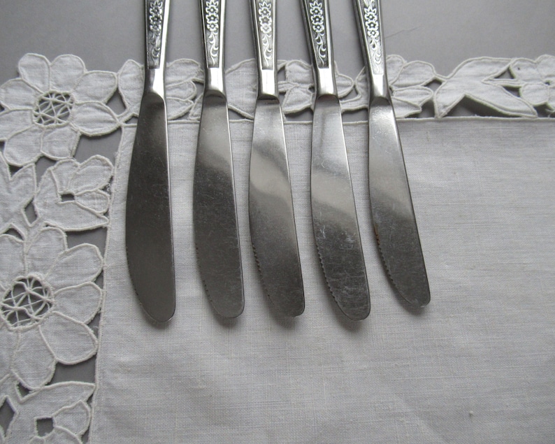 May include: Five silver butter knives with ornate handles lay on a white lace tablecloth.