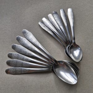 Könnte beinhalten: Ein Set aus zwölf silberfarbenen Metalllöffeln, die in zwei fächerartigen Gruppen angeordnet sind. Jeder Löffel hat einen langen Stiel mit einem geometrischen Muster aus Rauten und horizontalen Linien, das in einer glatten ovalen Laffe endet.