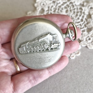 Può includere: Orologio da tasca argentato con un design a rilievo di un treno sul coperchio. L'orologio ha un'asta di carica e un anello per una catena. Il testo sull'orologio recita "Tram 70 years jubilee 1952-52".