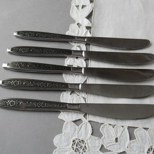 May include: Five silver dinner knives with floral designs on the handles. The knives are arranged in a row on a white lace tablecloth.