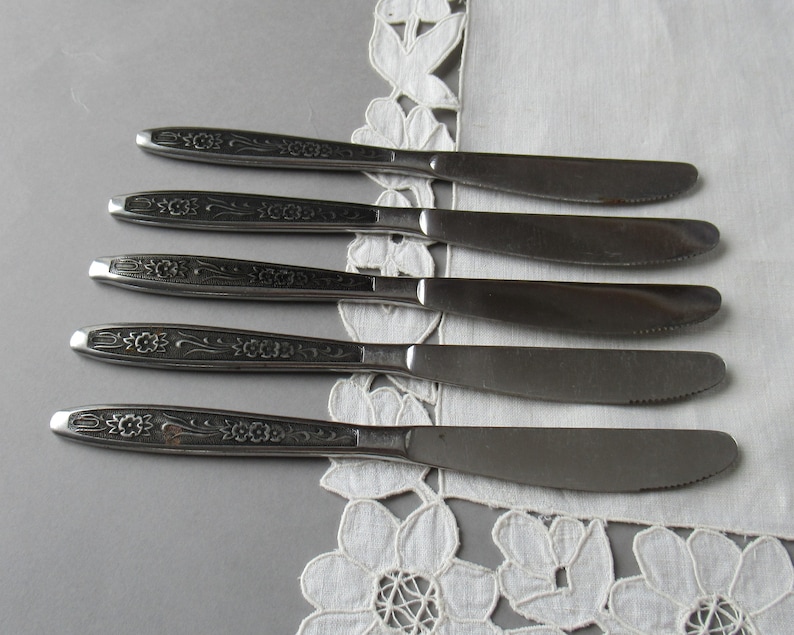 May include: Five silver dinner knives with floral designs on the handles. The knives are arranged in a row on a white lace tablecloth.