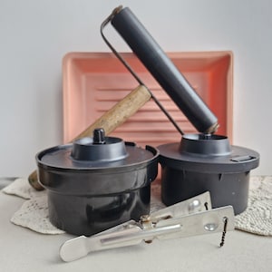 Może przedstawiać: Kolekcja zabytkowego sprzętu do ciemni, w tym dwa czarne zbiorniki do wywoływania filmów, szpula filmowa, klips do filmu i różowa tacka do wywoływania. Szpula filmowa ma drewnianą rączkę. Przedmioty ułożone są na białej powierzchni.