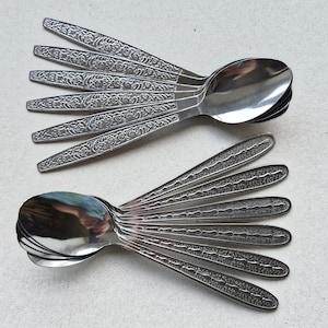 May include: A collection of twelve silver-toned metal spoons arranged in two groups. The top six spoons feature an intricate embossed floral pattern on the handles, while the bottom six spoons display a textured, dotted design along the stems.