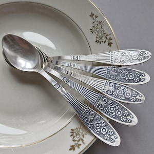May include: Five silver spoons with detailed floral and scroll patterns on the handles, arranged on a white plate with a gold rim and floral decoration. The spoons have a polished, reflective finish, and are made of silver.