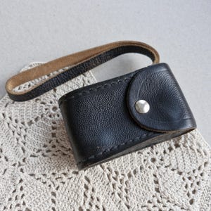 May include: A black leather case with a textured surface and a silver button closure. It has a brown leather strap attached. The case is resting on a cream-colored, crocheted doily.