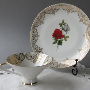 May include: A white teacup and saucer set with gold trim and a floral design. The plate features a red and white rose, while the cup and plate have gold filigree patterns. The set is displayed on a white surface.