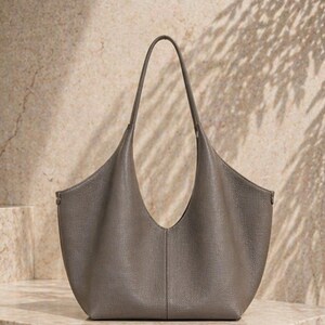 May include: A gray leather tote bag with a curved design and a single handle. The bag has a spacious interior and a minimalist aesthetic, suitable for everyday use. The bag is made of a soft, textured leather.