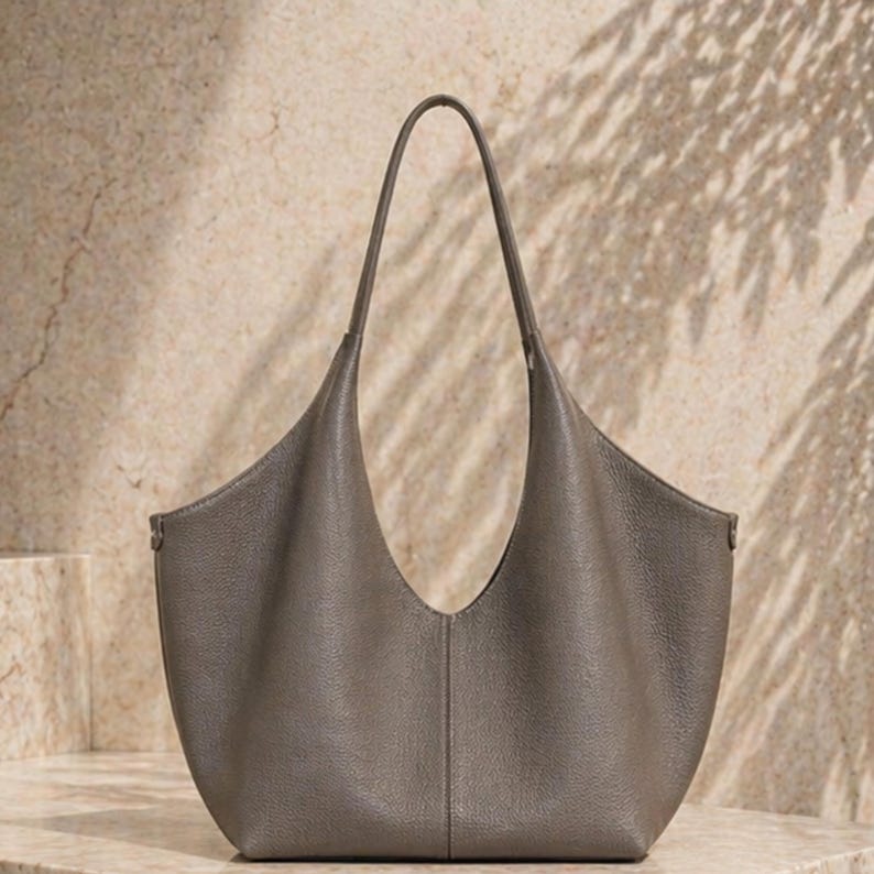 May include: A gray leather tote bag with a curved design and a single handle. The bag has a spacious interior and a minimalist aesthetic, suitable for everyday use. The bag is made of a soft, textured leather.