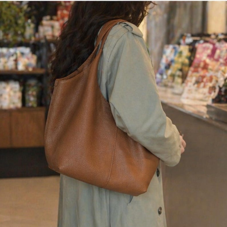 May include: A brown leather shoulder bag with a curved design. The bag has a single shoulder strap and appears to be empty. The bag is being carried by a person wearing a light green coat.
