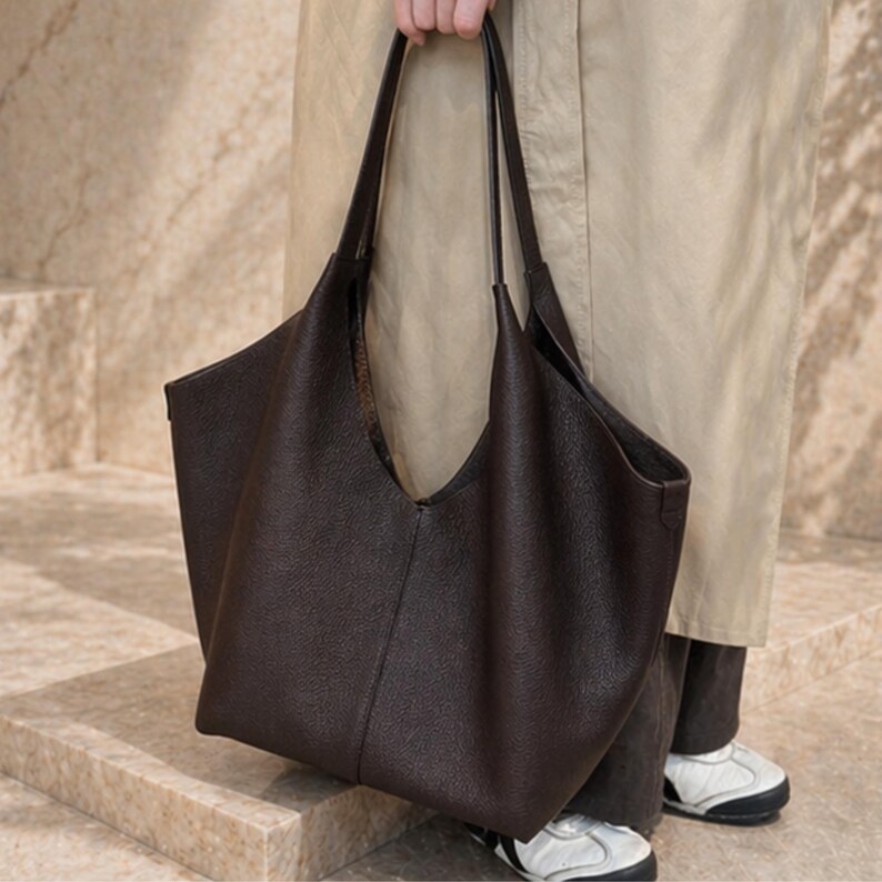 May include: A dark brown leather tote bag with long handles. The bag has a wide, open top and a textured surface. The bag is being held by a person wearing a tan coat and dark pants. White and black sneakers are visible.
