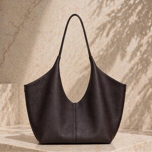 May include: A dark brown leather tote bag with a curved, open top and long, thin handles. The bag has a textured surface and a minimalist design, suitable for everyday use.