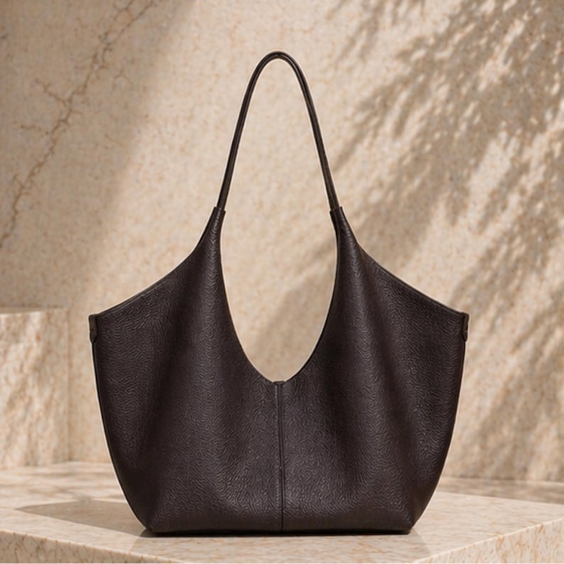 May include: A dark brown leather tote bag with a curved, open top and long, thin handles. The bag has a textured surface and a minimalist design, suitable for everyday use.
