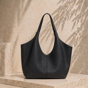 May include: A black leather tote bag with a unique, curved opening and two top handles. The bag has a textured surface and a minimalist design. The bag is sitting on a marble surface, with shadows in the background.