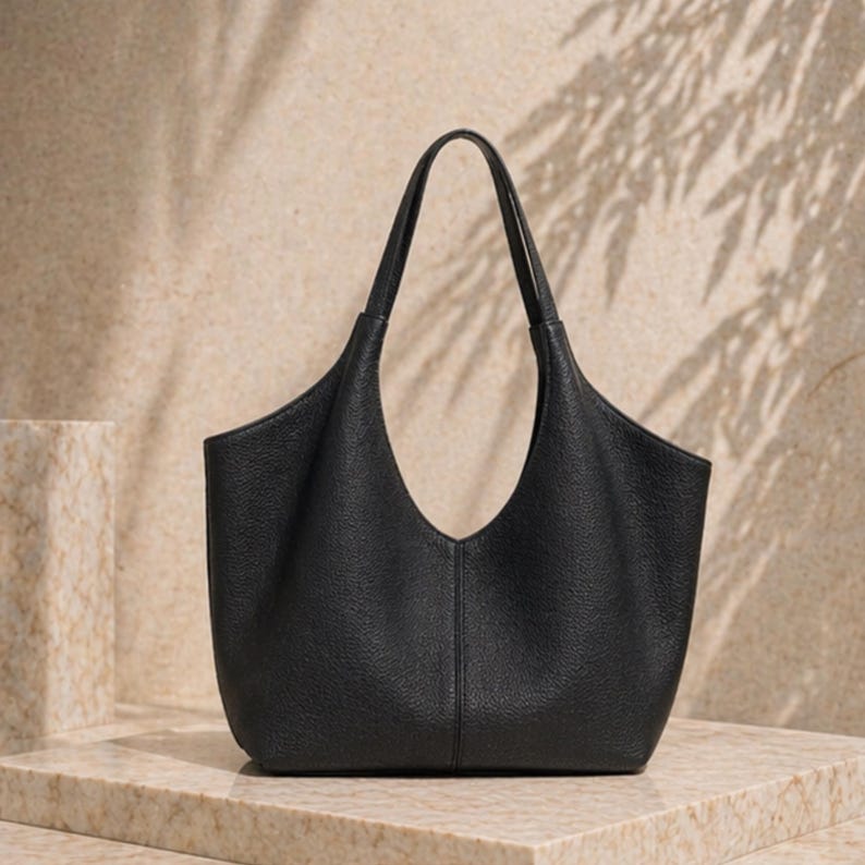 May include: A black leather tote bag with a unique, curved opening and two top handles. The bag has a textured surface and a minimalist design. The bag is sitting on a marble surface, with shadows in the background.