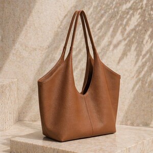 May include: A brown leather tote bag with long handles. The bag has a wide opening and a simple, elegant design. The bag is sitting on a stone surface.