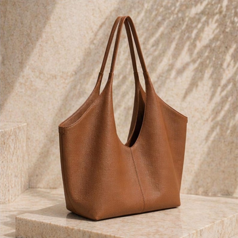 May include: A brown leather tote bag with long handles. The bag has a wide opening and a simple, elegant design. The bag is sitting on a stone surface.