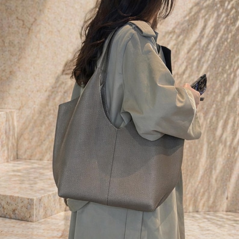 May include: A large, gray tote bag with a textured surface. The bag has a wide opening and a single shoulder strap. The bag is being carried by a person wearing a light green coat.