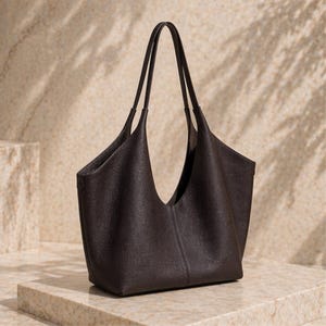 May include: A dark brown leather tote bag with a wide, open top and long handles. The bag has a structured shape and appears to be made of a textured leather material. The handles are thin and elegant.