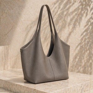 May include: A gray leather tote bag with long handles. The bag has a wide, open top and a structured shape. The bag is sitting on a marble surface. The background has a neutral color.