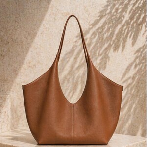 May include: A brown leather tote bag with a curved opening and long handles. The bag has a minimalist design and a smooth texture. The handles are thin and extend upwards. The bag is sitting on a light-colored surface.