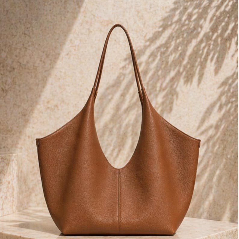 May include: A brown leather tote bag with a curved opening and long handles. The bag has a minimalist design and a smooth texture. The handles are thin and extend upwards. The bag is sitting on a light-colored surface.