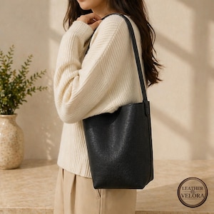 May include: Black leather tote bag with a shoulder strap. The bag has a textured surface and a simple, minimalist design. The bag is being held by a person wearing a cream-colored sweater.