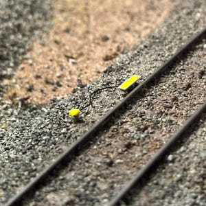 Könnte beinhalten: Eine Miniatur-Modelleisenbahnszene mit zwei parallelen Gleisen, einem gelben rechteckigen Licht und einer kleineren gelben Komponente, die durch einen schwarzen Draht verbunden sind. Die Szene befindet sich auf einem Bett aus kleinen grauen und braunen Steinen, das eine realistische Bahnumgebung simuliert.